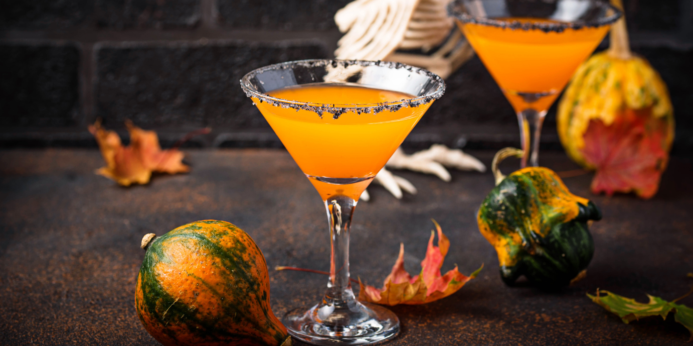 two martini glasses filled with orange liquid next to pumpkins and leaves