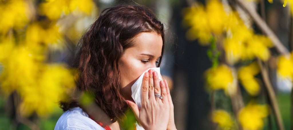 Can Seasonal Allergies Cause Migraines? What’s the Connection? a woman blowing her nose in front of a tree with yellow flowers